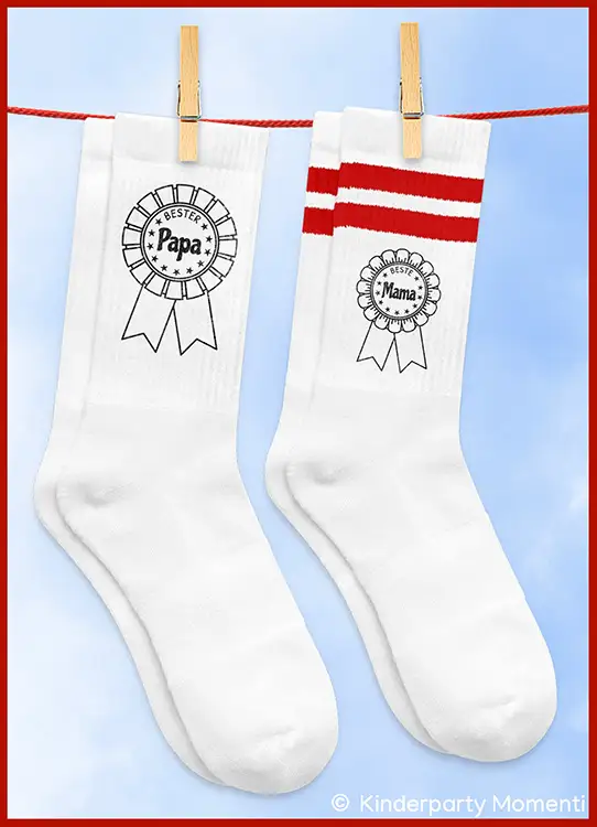 Zwei weiße Socken mit Aufdruck, eine mit rotem Streifenmuster und der Schrift 'Beste Mama', die andere mit der Schrift 'Bester Papa', beide an einer Wäscheleine befestigt