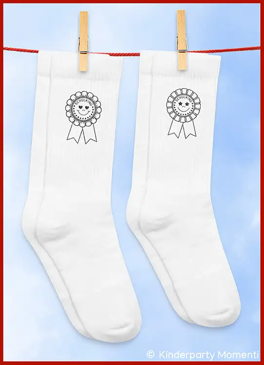 Zwei weiße Socken mit aufgedruckten Siegerrosen mit dem Text 'Schulkind' hängen an einer Wäscheleine vor blauem Himmel