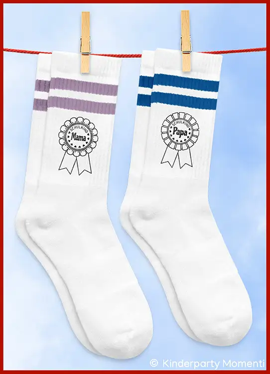 Zwei weiße Socken mit farbigen Streifen oben und den Aufdrucken 'Schulkind Mama' und 'Schulkind Papa' hängen an einer Wäscheleine