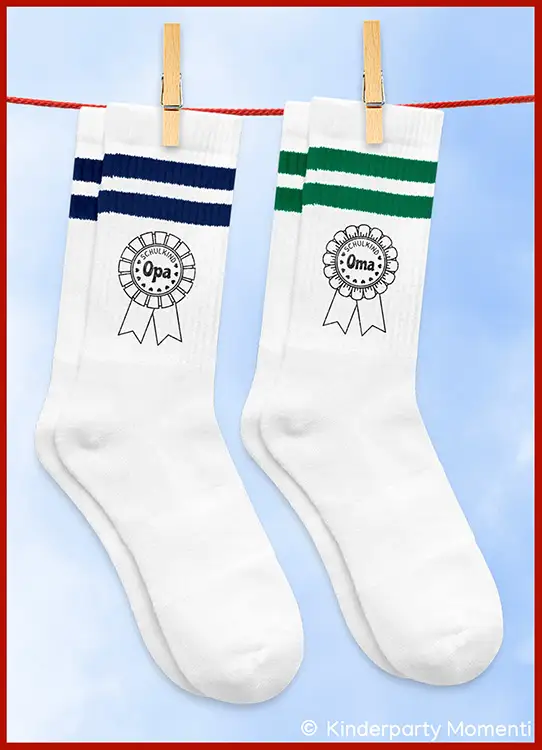 Zwei weiße Sportsocken mit je zwei Streifen oben, eine mit blauen Streifen und der Aufschrift 'Schulkind Opa', die andere mit grünen Streifen und der Aufschrift 'Schulkind Oma', an einer Wäscheleine mit Wäscheklammern aufgehängt