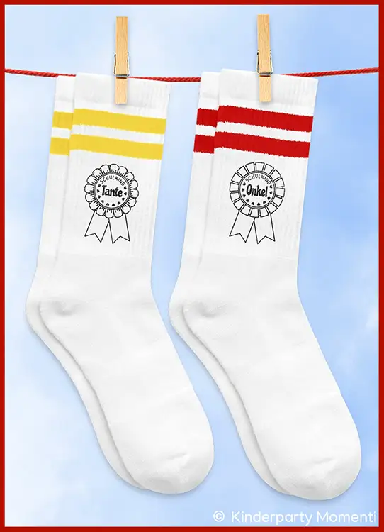 Zwei weiße Socken mit gelben und roten Streifen oben, jeweils mit einer Orden-Schleife und den Worten 'Schulkind Tante' und 'Schulkind Onkel' darauf, an einer Wäscheleine befestigt