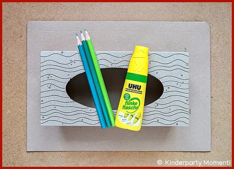 Drei Buntstifte in Blau- und Grüntönen, eine gelbe UHU-Klebestiftflasche und eine Taschentuchbox mit Wellenmuster und ovalem Ausschnitt auf braunem Untergrund