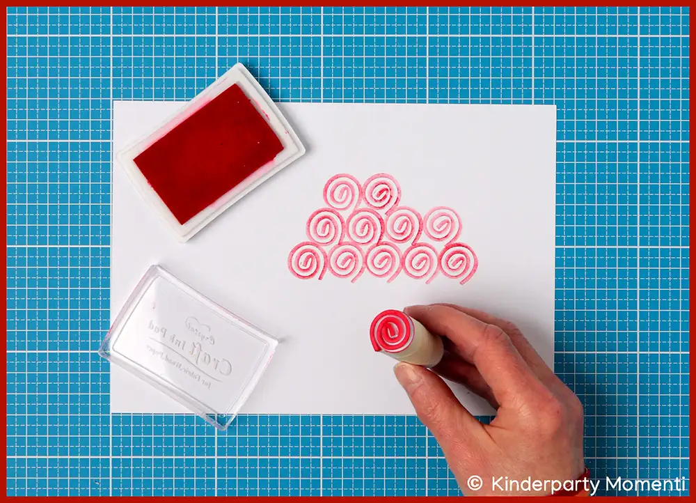 Hand stempelt mit rotem Moosgummistempel Spiralmuster auf weißes Papier neben rotem Stempelkissen auf blauem Schneideunterlage