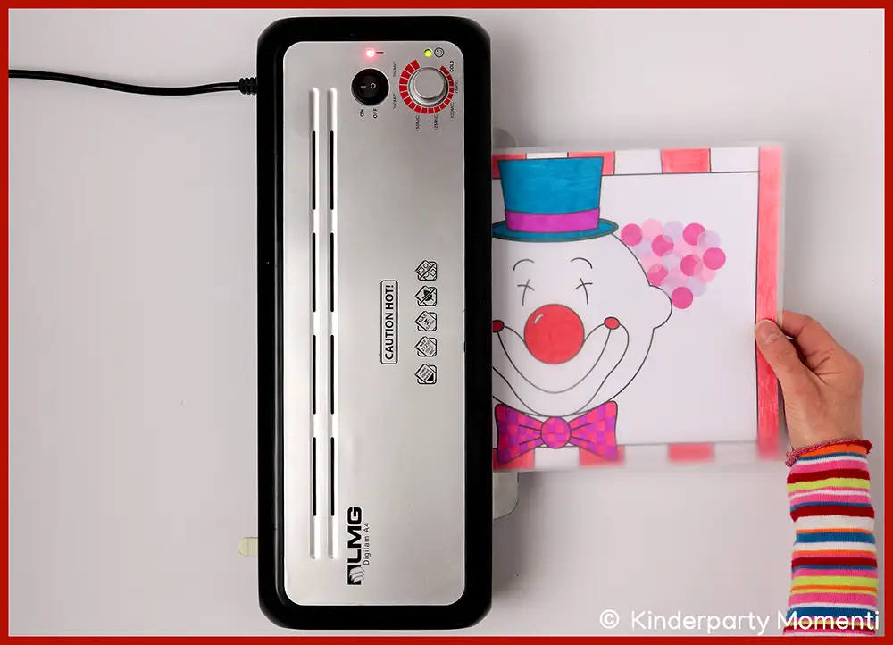 Laminiergerät , das ein buntes Bild mit Clown Gesicht laminiert, gehalten von einer Hand in buntem Ringelshirt