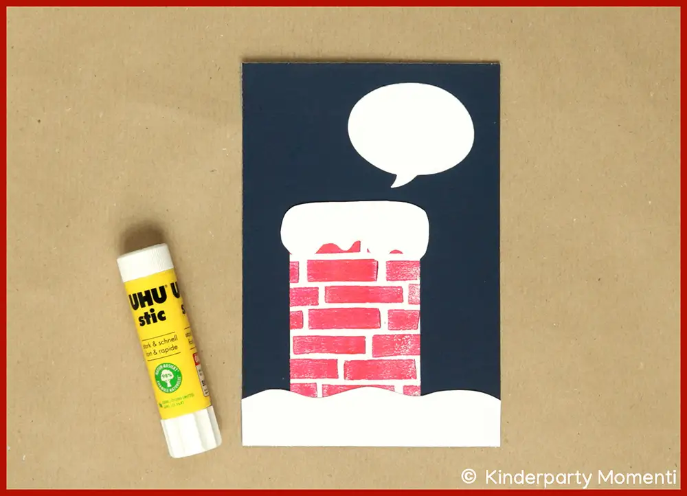 DIY Weihnachstskarte mit rotem Schornstein, weißem Schnee und leerer Sprechblase auf dunkelblauem Hintergrund, daneben ein gelber UHU-Klebestift.