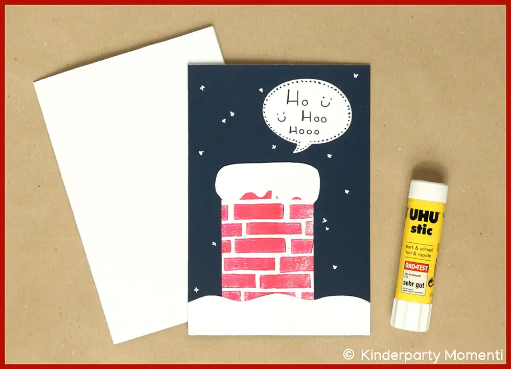 Selbstgestaltete Weihnachtskarte mit rotem Ziegel-Schornstein und Sprechblase 'Ho Ho Hooo' neben weißem Kartenrohling und UHU-Stic-Klebestift.