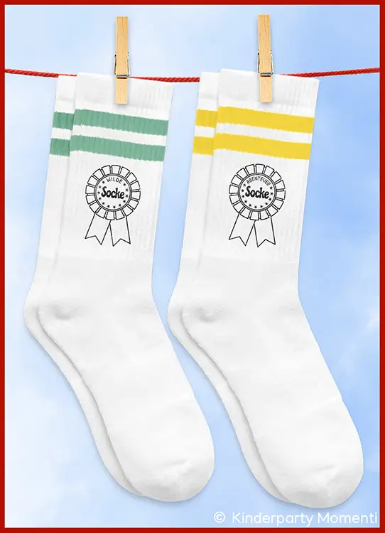 Zwei weiße Socken mit je zwei Streifen oben, eine grün, eine gelb, beide mit einem Aufdruck eines Ordens mit dem Wort 'Wilde Socke' und 'Abenteuer Socke', an einer roten Leine mit Wäscheklammern hängend