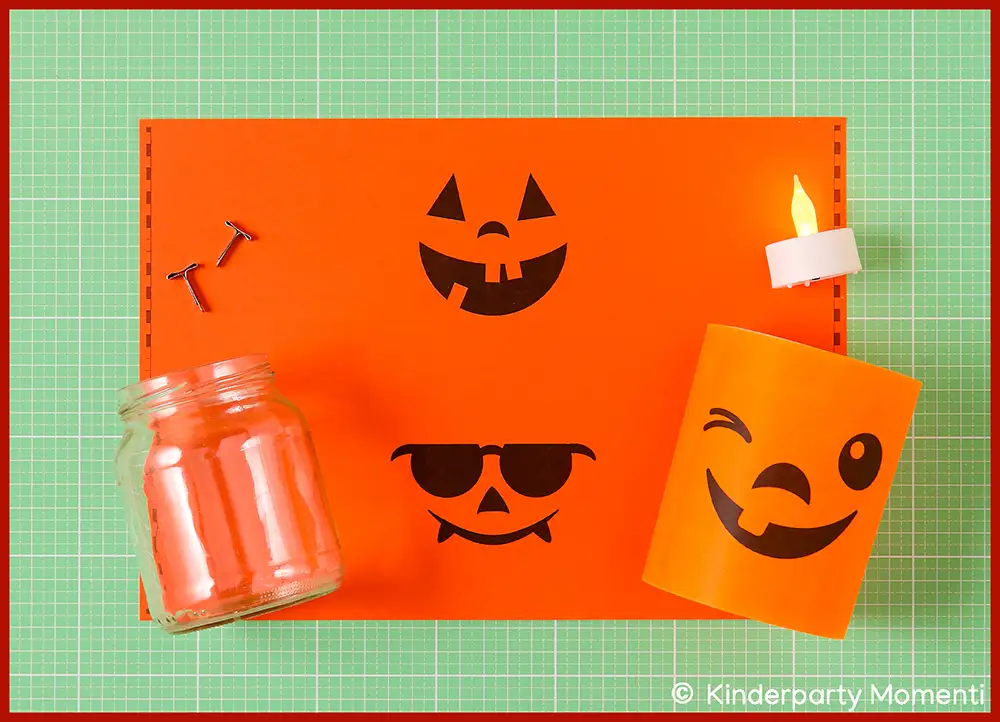 Ausdruckbare Vorlage mit schwarzen Halloween Kürbisgesichtern, ein leeres Glas, zwei Musterklammern und eine LED-Kerze auf einer grünen Schneidematte