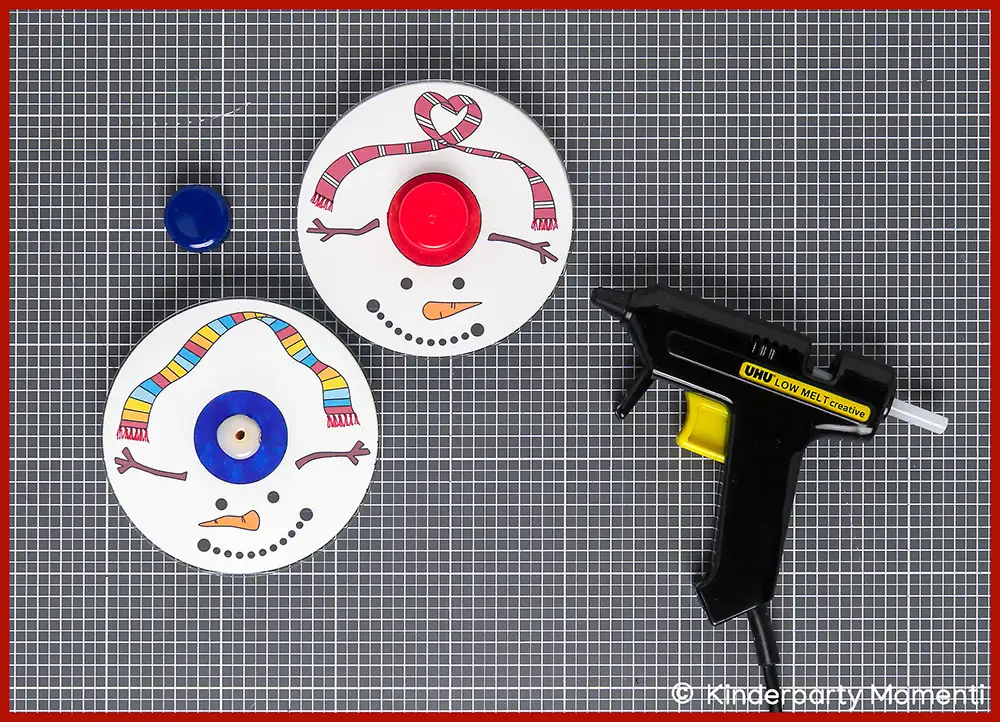 03 Kreisel Basteln aus CDs - Flaschendeckel als Drehknopf auf die CD kleben Zwei mit Schneemanngesichtern bemalte CDs und eine schwarze Heißklebepistole auf grauer Bastelmatte