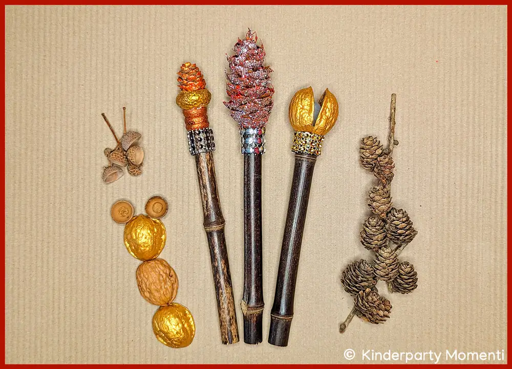 Hexen Zauberstäbe selbst machen mit Naturmaterial Drei DIY Zauberstäbe mit verzierten Enden aus gold- und rotfarbenen Nüssen und Zapfen, daneben mehrere goldene und natürliche Nüsse sowie ein Zweig mit kleinen Zapfen auf braunem Karton.