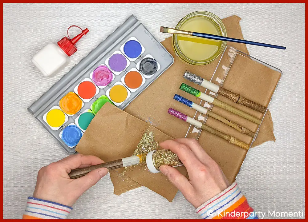 Hexen Zauberstab selber machen Glitzer-Steinen & Glitter Hände halten Pinsel und Becher mit goldfarbenem Glitter über einen Holzstab, daneben Tuschkasten, Kleberflasche, Pinsel und selbstgebastelte Glitzer Zauberstäbe