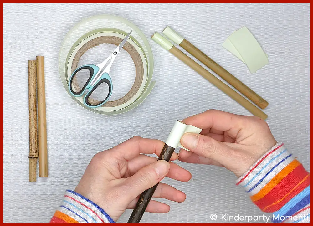 Nachleuchtenden Hexen Zauberstab selber machen mit Kindern Hände kleben Glow-in-the-Dark Klebeband auf Bambus Holzstab, daneben Schere, nachleuchtendes Klebeband und zwei DIY Zauberstäbe