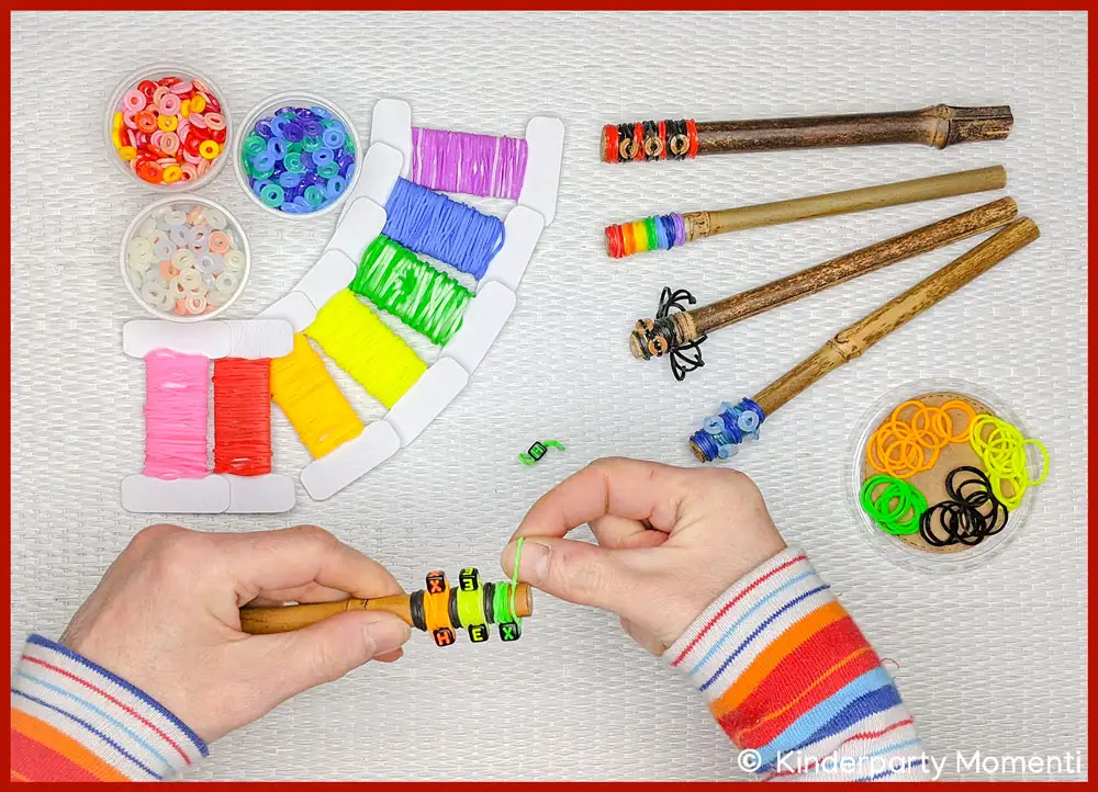 Hexen Zauberstab selbst machen mit Perlen und Loom Gummibändern Hände fädeln Loom Gummis und Perlen auf Holzstab, daneben Becher mit Perlen, Loom Bänder und vier gebastelte Zauberstäbe