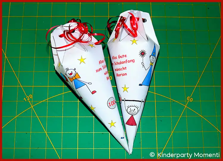 Zwei Schultüten aus Papier mit bunten Clownzeichnungen, Sternen und roter Schleife auf grünem Schneideunterlage mit Raster